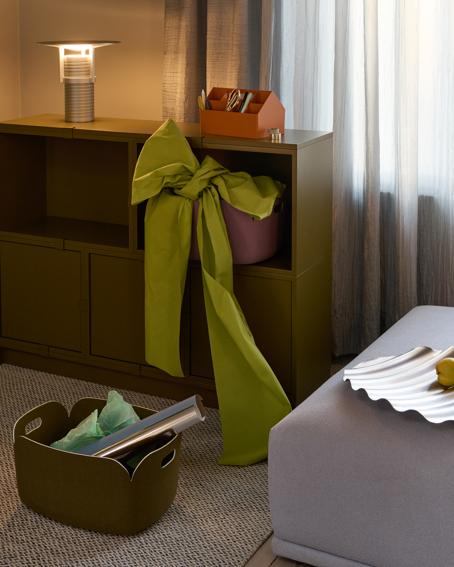 Image of a Muuto Restore Basket in dark green, featuring a large bow in the center to enhance the holiday atmosphere. The scene also includes a Set Table Lamp, a Wave Tray, and a Sketch Toolbox, all displayed in a cozy holiday 2024 decor setting.